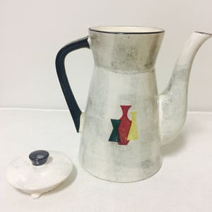 May include: White ceramic coffee pot with a black handle and a lid. The pot has a geometric design of three stylized vases in green, red, and yellow.
