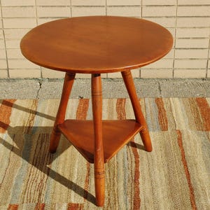 May include: A round wooden table with a triangular shelf. The table has three legs and is made of a light brown wood.