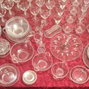 Puede incluir: Una gran colecci&oacute;n de cuencos, platos y piezas de servir de vidrio transparente con bordes perlados. Las piezas est&aacute;n dispuestas sobre un mantel rojo con un patr&oacute;n perlado.