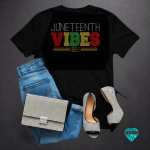 May include: A black t-shirt with the words "Juneteenth Vibes" in red, yellow, and green letters. The year "1865" is printed below the text. The shirt is displayed on a model with blue jeans, a silver purse, and silver high heels.