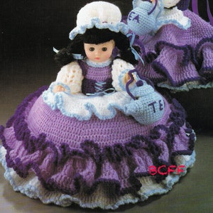 May include: Crocheted doll dress with a purple and white skirt, a white bodice, and a white hat. The doll is holding a crocheted teapot that says "Tea".