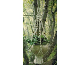 May include: A white macrame plant hanger with a green plant in a brown pot. The hanger is hanging from a tree branch in a forest setting.