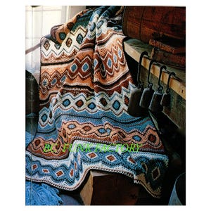 Peut inclure: Une couverture en crochet présentant un motif géométrique répétitif dans des tons de bleu, marron, crème et blanc. Le textile affiche des motifs complexes en losange et en zigzag sur toute sa surface. Le texte "BC FUNK FACTORY" est en vert.