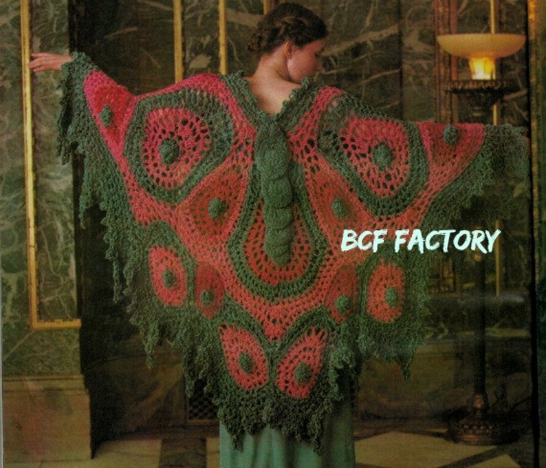 May include: A green and pink crocheted butterfly-shaped shawl with a textured design. The shawl is worn by a person standing in front of a patterned wall. BCF FACTORY is printed on the shawl.
