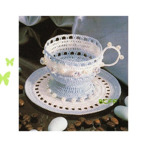 May include: A crocheted teacup and saucer set in blue and white with pearl accents. The set is sitting on a surface with coffee beans.