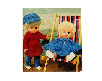 Op de afbeelding: Twee poppen gekleed in gebreide kleding. De pop links draagt een rode gebreide jurk, een blauwe broek en een blauwe hoed. De pop rechts draagt een blauwe gebreide cape en zit in een rood-wit-blauwe gestreepte strandstoel.