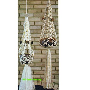 Op de afbeelding: Twee macramé plantenhangers. Eén is crèmekleurig met vlinderaccenten en een kwast. De andere is bruin met een bruine pot en een lange kwast. Beide hangen tegen een bakstenen muur.