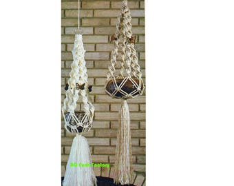 May include: Two macrame plant hangers. One is cream-colored with butterfly accents and a tassel. The other is tan with a brown pot and a long tassel. Both are suspended against a brick wall.