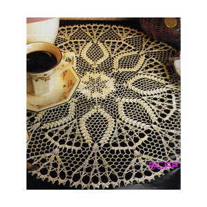 May include: A delicate, off-white crocheted doily with a floral design. The doily is circular and has a lace-like pattern. It is placed on a dark surface with a cup of coffee and a saucer.