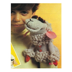 Pode incluir: Um fantoche de meia de lã cinza e branca em forma de ovelha. O fantoche tem um nariz rosa e olhos pretos. O fantoche está sendo segurado por uma criança.