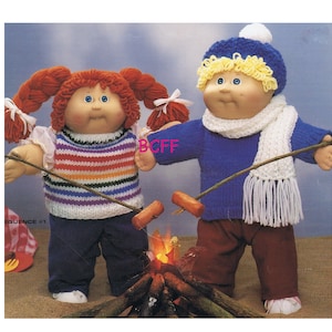May include: Two Cabbage Patch Kids dolls dressed in knitted clothing. The girl doll has red hair and is wearing a rainbow striped vest and blue jeans. The boy doll has blonde hair and is wearing a blue sweater, white scarf, and brown trousers. Both dolls are holding sticks with hot dogs on them, and there is a small fire in the background.