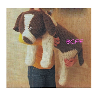May include: A crocheted stuffed dog toy in brown and white with black eyes. The dog is being held by a person wearing a yellow shirt and blue jeans.