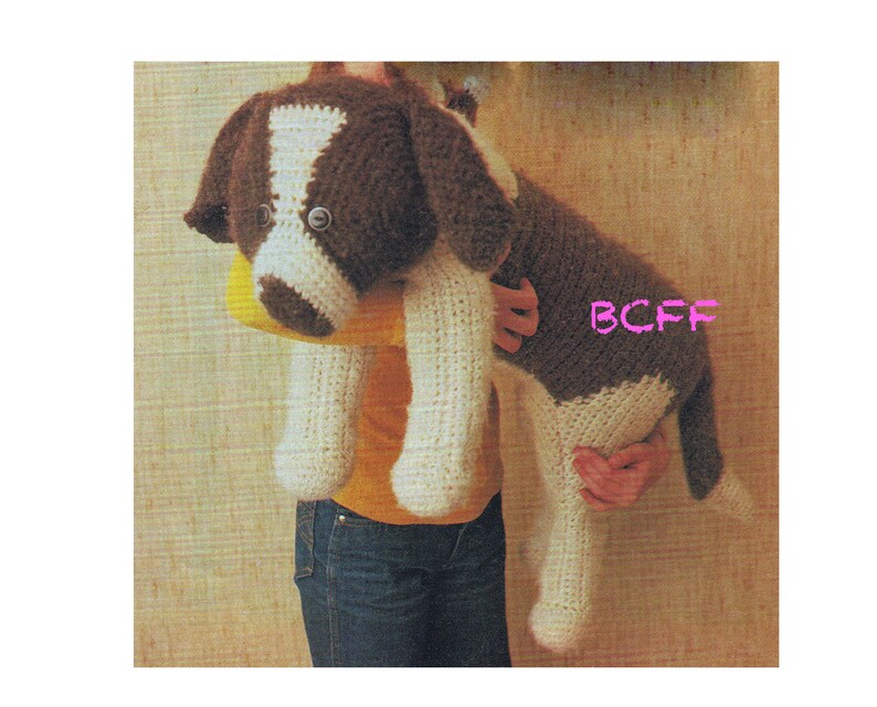 May include: A crocheted stuffed dog toy in brown and white with black eyes. The dog is being held by a person wearing a yellow shirt and blue jeans.