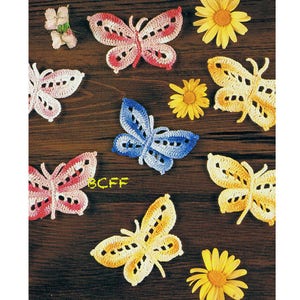 May include: A collection of seven crocheted butterflies in various colours, including pink, blue, yellow, and white. The butterflies are arranged on a wooden surface with a few small flowers.