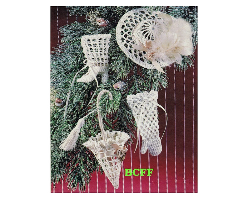 Op de afbeelding: Witte gehaakte kerstversieringen in de vorm van een laars, een hoed, een paraplu en een kous. De versieringen hangen aan een groene kerstboom.