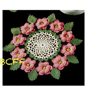 Könnte beinhalten: Eine weiße Häkeldeckchen mit einem floralen Design. Das Deckchen ist von acht rosa gehäkelten Blumen mit grünen Blättern umgeben.