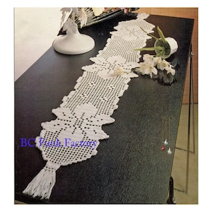 Puede incluir: Un camino de mesa de crochet blanco con un diseño floral y una borla en el extremo. El camino de mesa está sobre una mesa negra con un jarrón blanco y flores.