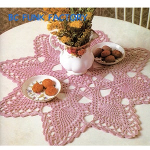 May include: A pink crocheted doily with a starburst design. The doily is on a white table with a vase of flowers and a small plate of cookies.