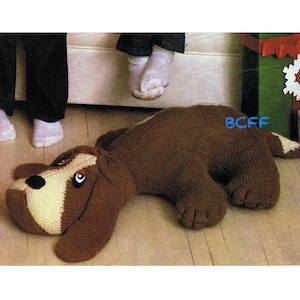 Może przedstawiać: Pluszowa zabawka w kształcie psa w kolorze brązowym i kremowym leżąca na drewnianej podłodze. Pies ma długie ciało i krótki ogon. Oczy psa są czarne, a nos brązowy.