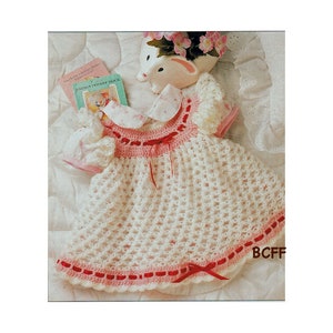 Peut inclure: Une robe de bébé en crochet blanc avec une bordure rose et rouge. La robe a un petit nœud rose à la taille et un col en crochet blanc.