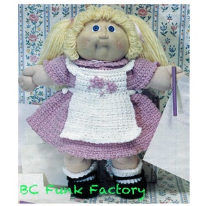 May include: A Cabbage Patch Kid doll wearing a pink dress with a white crocheted apron. The doll has blonde hair in pigtails and blue eyes. The doll is holding a purple pencil and a piece of paper.