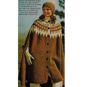 May include: A brown knitted cape with white Scandinavian designs. The cape has a high collar and is fastened with buttons down the front. The model is wearing a white knitted hat.