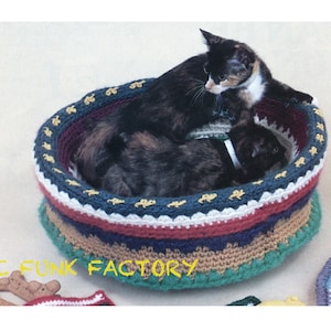 May include: Two cats resting in a colorful crocheted cat bed. The bed is made with a variety of colors, including red, blue, green, and brown. The bed is round and has a raised edge. The cats are both black and white.