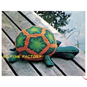 Peut inclure: Un jouet en crochet en forme de tortue avec une carapace verte et des accents orange. La tortue est posée sur une surface en bois.