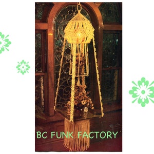 Peut inclure: Un porte-plante en macramé de couleur crème avec une étagère carrée en verre et une base à franges, suspendu devant une fenêtre. Le texte "BC FUNK FACTORY" est en bas.