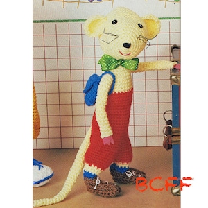 Puede incluir: Una muñeca de ratón de crochet que lleva un mono rojo, una mochila azul y una pajarita verde. El ratón está de pie junto a una superficie a rayas azul y blanca.