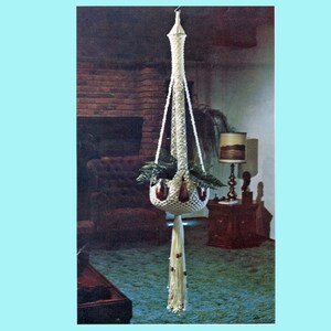 May include: A vintage macrame plant hanger, suspended from the ceiling. The white woven basket holds greenery and decorative brown beads. The long, fringed tail adds a bohemian touch. The background includes a brick fireplace, a lamp, and a dark armchair.