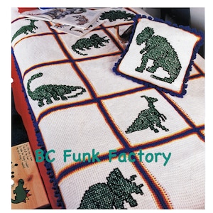May include: A white crocheted blanket with a blue and orange border features a variety of green dinosaur designs. The blanket is decorated with blue pom-poms. A matching pillow with a green dinosaur design is also included. The text "BC Funk Factory" is visible in the image.