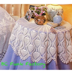 Puede incluir: Un mantel de mesa de crochet blanco con un borde festoneado y un diseño de piña. El mantel está sobre una mesa azul claro con un jarrón de flores rosas y una botella decorativa.
