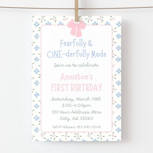 May include: A rectangular invitation card with a pink bow at the top. The card has a floral pattern border with blue flowers. The text reads "Fearfully & ONE-derfully Made" and announces "Anniston's FIRST BIRTHDAY" on March 15th.