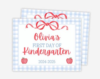 May include: Two stacked, rectangular signs with a light blue gingham border. The top of each sign features a red bow. The text reads "Olivia's First Day of Kindergarten 2024-2025" in red and blue. Red apple illustrations are on each side.