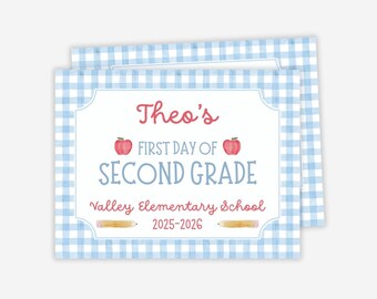 May include: A personalized school sign with a blue gingham border. The sign reads "Theo's First Day of Second Grade" with red apple illustrations and the school name. The year 2025-2026 is also included.