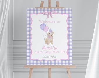 May include: A watercolor-style birthday sign on an easel. The sign features a light brown puppy wearing a party hat and holding balloons. The sign reads "Welcome to Lexi's 2nd birthday PAW-TY!" in pink text, with a purple and white gingham border.