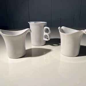 Może przedstawiać: Trzy białe ceramiczne dzbanki i kubek ustawione na białej powierzchni. Dzbanki mają unikalne, zakrzywione wylewki i uchwyty. Kubek ma charakterystyczny uchwyt z dwoma okrągłymi otworami. Obiekty rzucają cienie.
