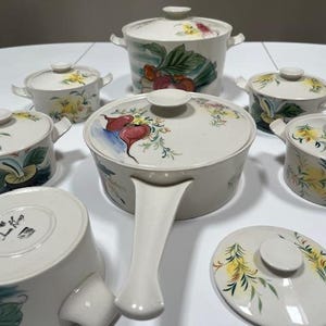 May include: A set of seven white ceramic cookware with floral and fruit designs. The set includes a large pot with a handle and six smaller bowls with lids.