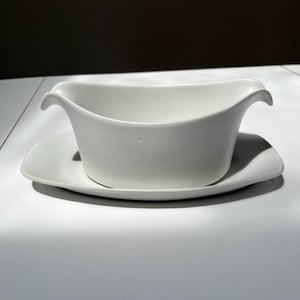 Może przedstawiać: Biała ceramiczna łódka do sosu z pasującym prostokątnym spodkiem. Łódka do sosu ma zakrzywiony uchwyt po każdej stronie i dzióbek do nalewania. Zestaw wykonany jest z gładkiego, błyszczącego materiału.