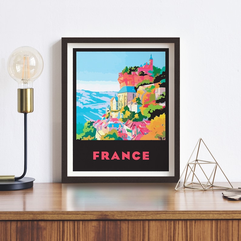 May include: A framed poster with a colorful illustration of a village in France. The village is built on a cliff with a church at the top. The poster has the word "FRANCE" in pink letters at the bottom.