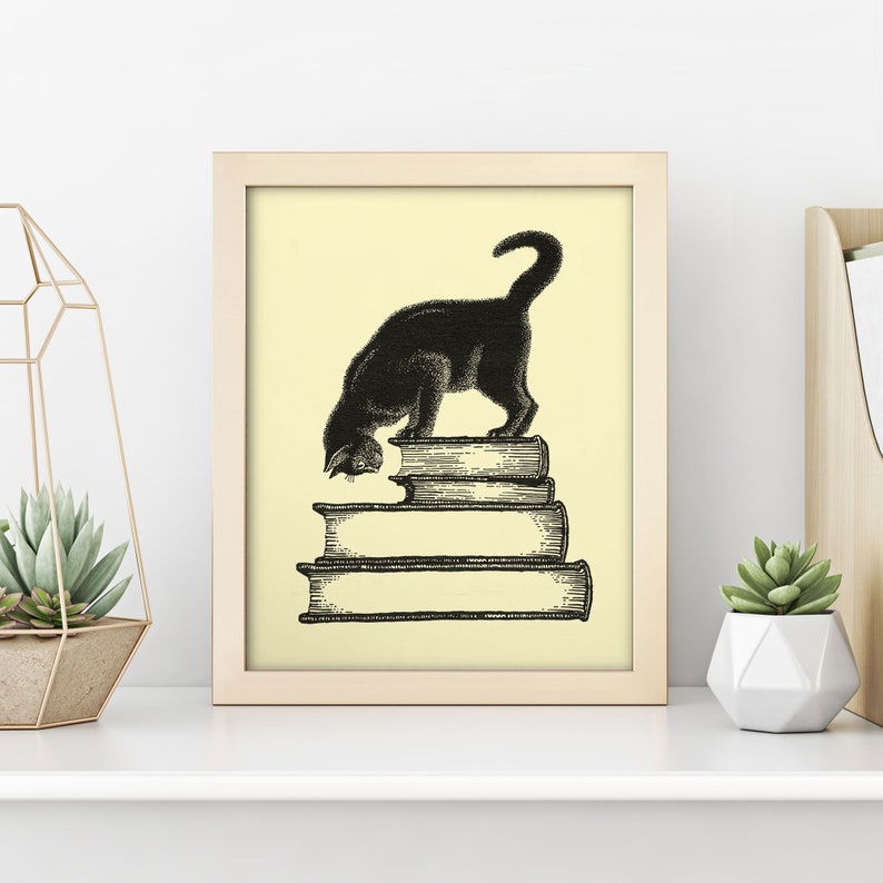 May include: A framed print featuring a black cat standing atop a stack of books. The cat is depicted in a vintage style, with its head lowered as if examining the books. The background is a soft yellow, and the frame is light wood.