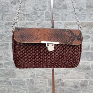 Può includere: Una borsa a mano marrone all'uncinetto con patta a motivo serpente e tracolla a catena argentata. La borsa ha una forma rettangolare e una chiusura con fermaglio argentato. La borsa è sospesa contro un muro di mattoni grigi.