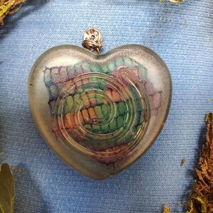 May include: A silver heart-shaped pendant with a swirling green, orange, and purple design inside. The pendant has a silver loop at the top for hanging.