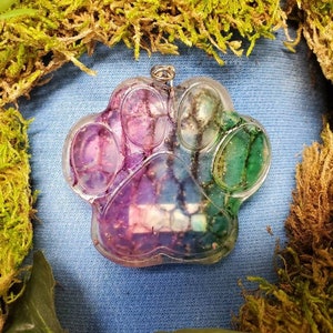 May include: A clear, paw-shaped pendant with a silver loop for hanging. The pendant is filled with a mix of purple, blue, and green glitter and resin. The background is a light blue fabric, surrounded by green moss.