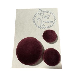 May include: Three burgundy fabric-covered buttons of varying sizes are arranged on a light-colored card. The card has a blue floral design with the word "enjoy" in script. The buttons have a soft, textured appearance.