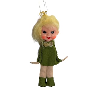 May include: A vintage Christmas ornament featuring a doll-like figure with blonde hair, rosy cheeks, and a red mouth. The doll wears a green felt dress and boots, with tan felt legs and arms. A gold bow adorns the dress.