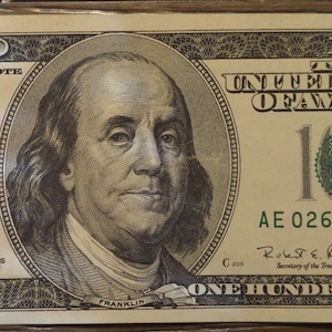 May include: A US $100 bill with a portrait of Benjamin Franklin. The bill has the serial number AE 02689479 and the text "THE UNITED STATES OF AMERICA" and "ONE HUNDRED DOLLARS".