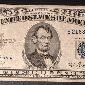 May include: A vintage five dollar silver certificate featuring a portrait of Abraham Lincoln. The certificate is dated 1953 and has the serial number E21884859A. The text "This certifies that there is on deposit in the Treasury of the United States of America" is printed on the top of the certificate.