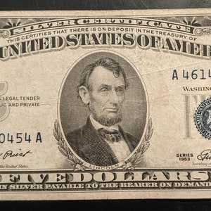 May include: A vintage five dollar bill featuring a portrait of Abraham Lincoln. The bill is printed in black and white with blue ink accents. The serial number is A 46140454 A. The text "THE UNITED STATES OF AMERICA" is printed at the top of the bill.